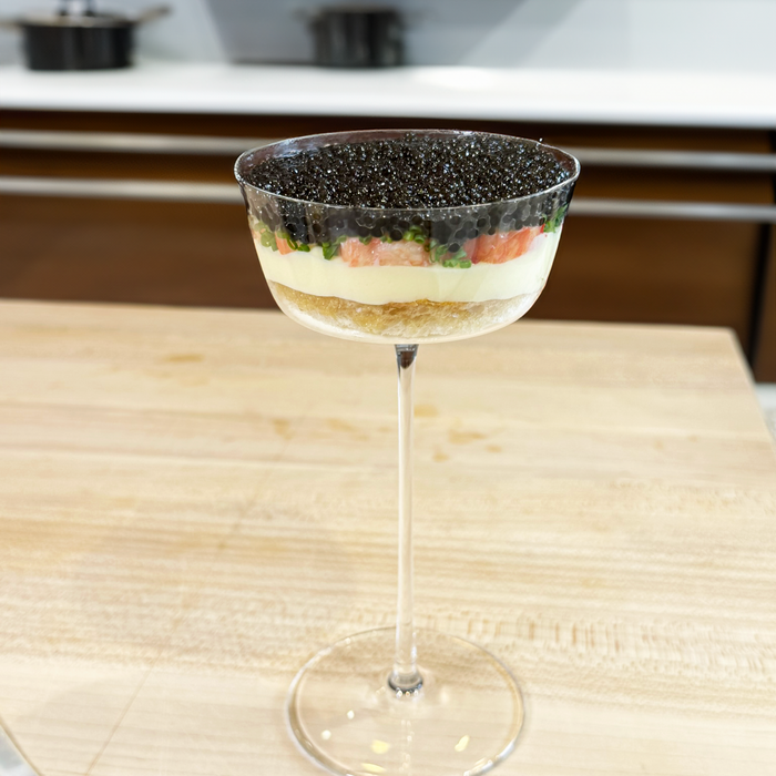 a thin glass with layers of bread, white potato, pink crab, and caviar on a wood cutting board