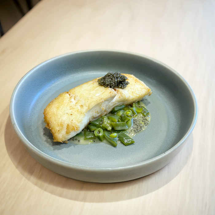 white fish on a bed of greens with a dollop of black caviar in a blue sage plate on a wood surface
