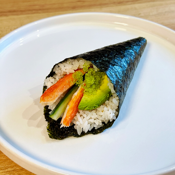 Close up of a dark green seaweed cone filled with rice, crabmeat, cucumber, avocado, and green tobikko on a white plate.