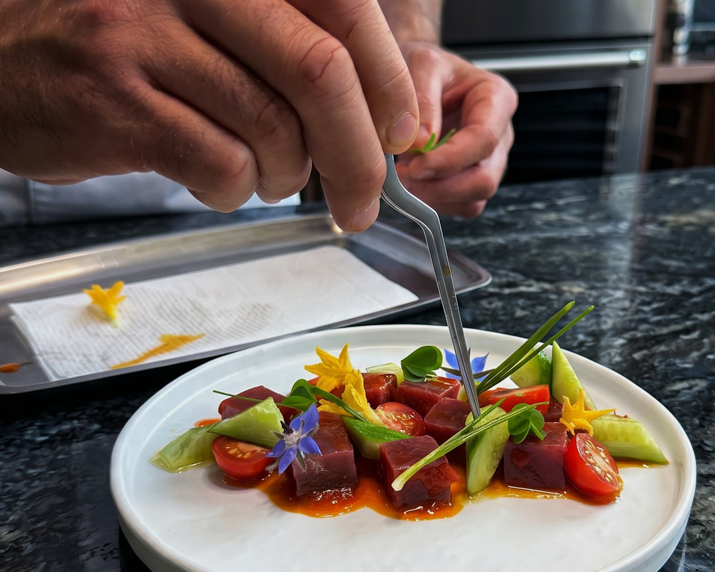 Person garnishing a colorful dish on a marble plate with a kitchen in the background