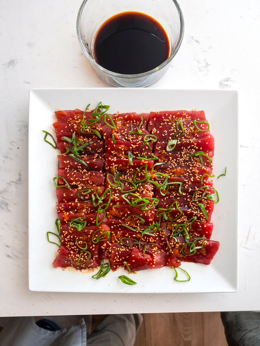 Raw tuna with sesame seeds and green onions on a white plate with a side of soy sauce.