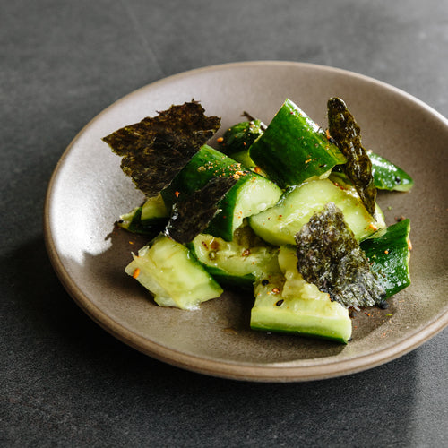 Chopped cucumber salad garnished with pieces of dark seaweed on a beige ceramic plate against a dark surface background.