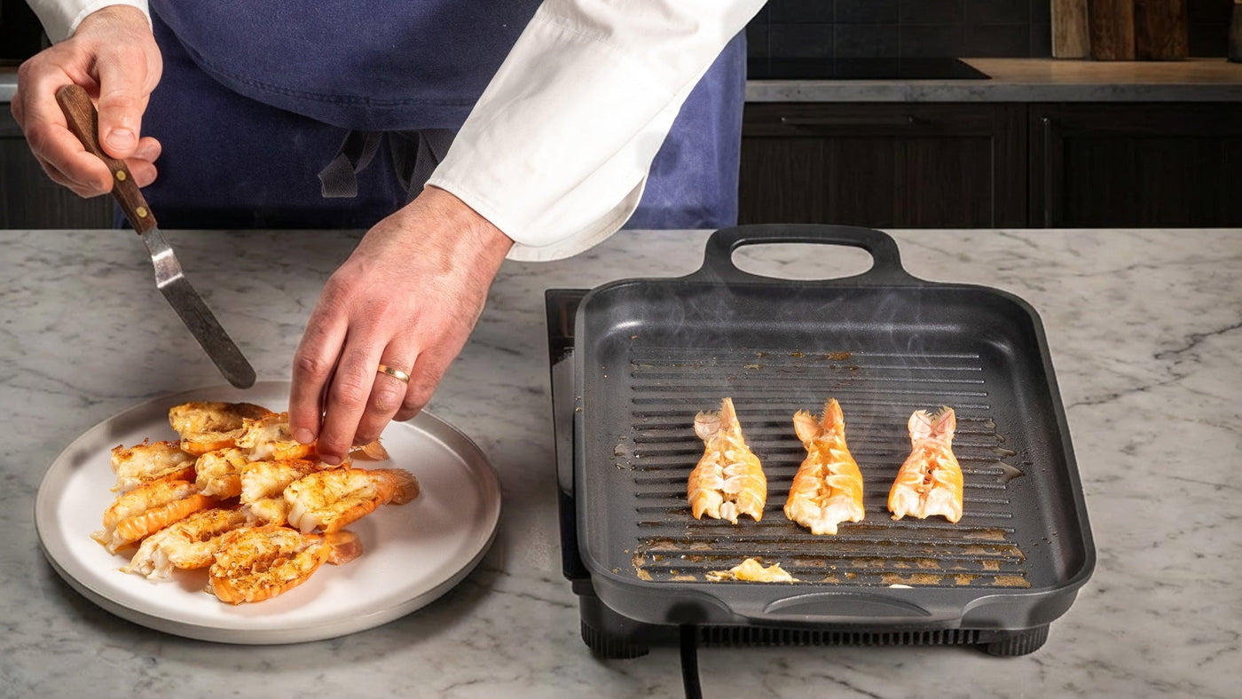Person grilling langoustine tails on a portable grill with a plate of food nearby in a kitchen setting.