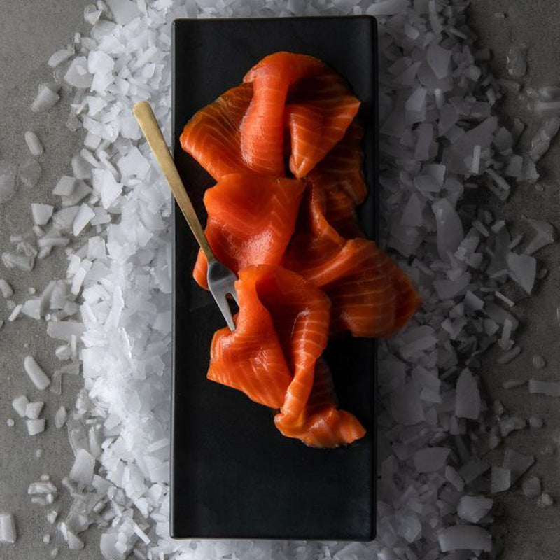 Thinly sliced vibrant orange Smoked Ora King Salmon artfully arranged on a black rectangular plate surrounded by crushed ice with a small gold fork.