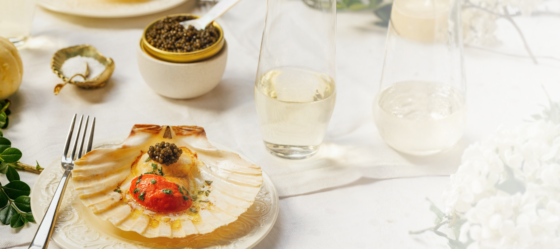 Delicate dish of a scallop topped with caviar, on a white tablecloth, accompanied by glasses of white wine.