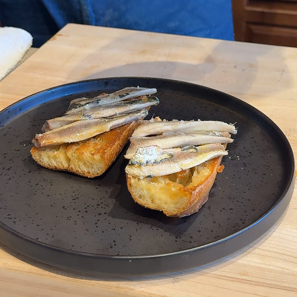 Two slices of toasted bread topped with sliced  grilled sardines, served on a dark speckled plate on a light wood table.