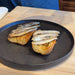 Two slices of toasted bread topped with sliced  grilled sardines, served on a dark speckled plate on a light wood table.