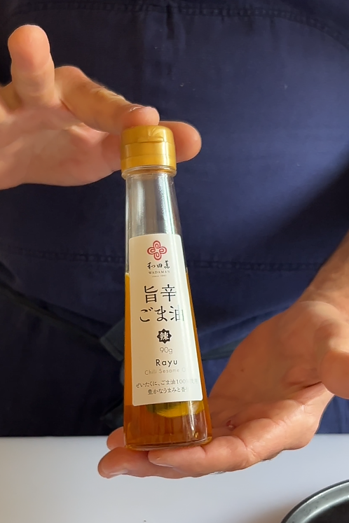 A person in a navy apron holds a small bottle of toasted sesame oil with a gold cap, showing the amber-colored liquid inside. A black plate with sliced food is partially visible on a white table.