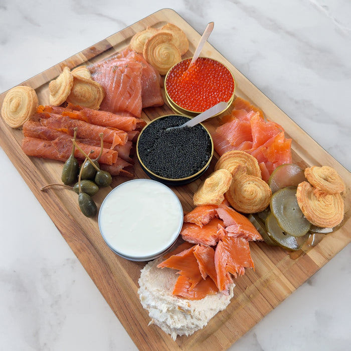 Wooden board with smoked salmon slices, smoked trout, two tins of trout roe and siberian caviar with spoons, creme fraiche, pickled vegetables, caper berries, and puff pastry crackers on a marble surface.