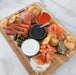Wooden board with smoked salmon slices, smoked trout, two tins of trout roe and siberian caviar with spoons, creme fraiche, pickled vegetables, caper berries, and puff pastry crackers on a marble surface.