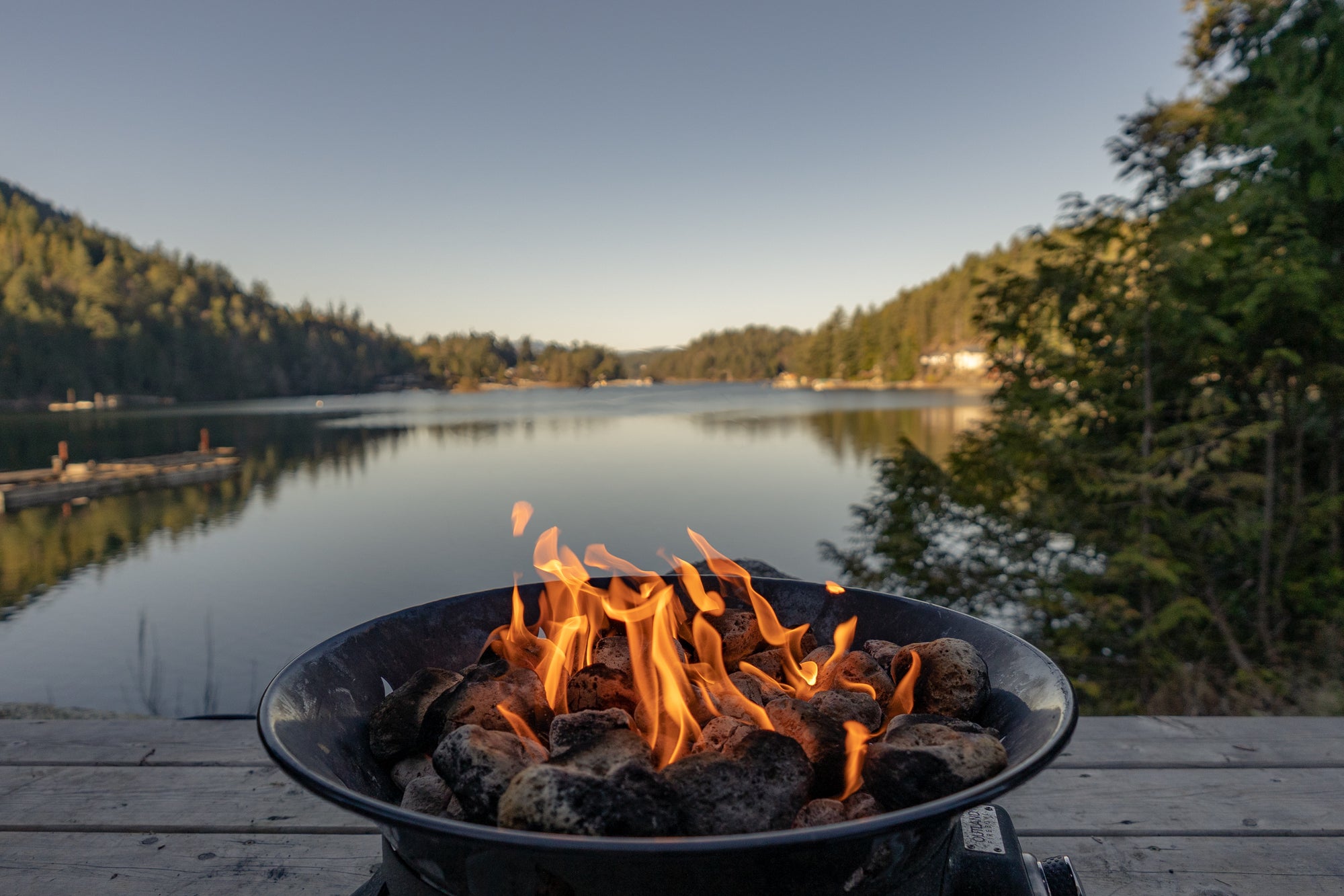 A fire pit with bright orange flames burning over charcoal, set on a wooden deck overlooking a calm lake surrounded by green trees under a clear sky.