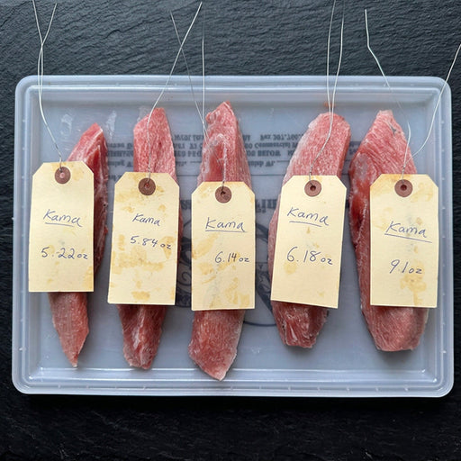 Five raw cuts of bluefin tuna collar "kama toro" set on a clear plastic tray, each with a handwritten paper tag labeled 'kama' and different weights, set against a dark slate background.