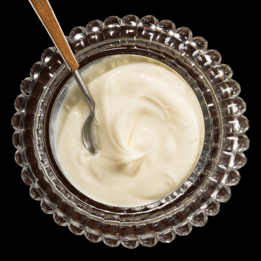 Top-down view of a clear glass bowl with a scalloped edge filled with smooth creme fraiche and a small spoon with a wooden handle resting inside.