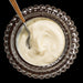 Top-down view of a clear glass bowl with a scalloped edge filled with smooth creme fraiche and a small spoon with a wooden handle resting inside.