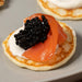 Close-up of a small golden blini topped with a dollop of creme fraiche, a slice of smoked salmon, and a generous scoop of Royal Belgian Siberian caviar 