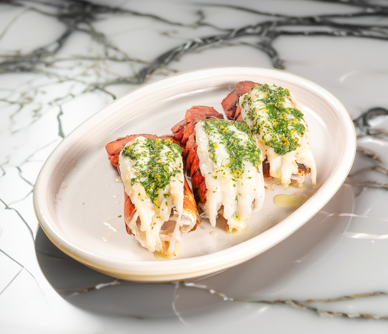 Three cooked lobster tails topped with herb butter sauce served on an oval white plate against a white marble surface background