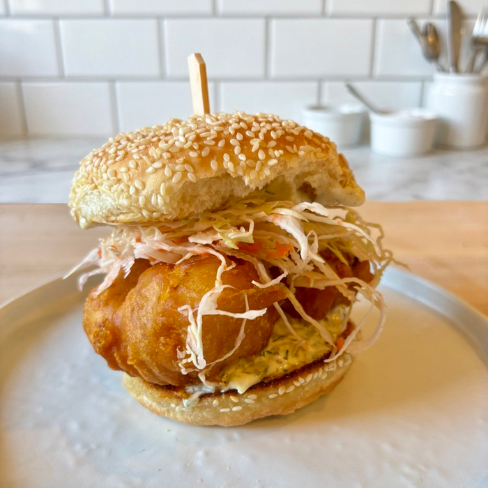 A close-up shot of a gourmet fish and chip sandwich on a white plate, featuring a golden-fried fish patty, shredded cabbage, and a sesame bun, set against a blurred kitchen background.