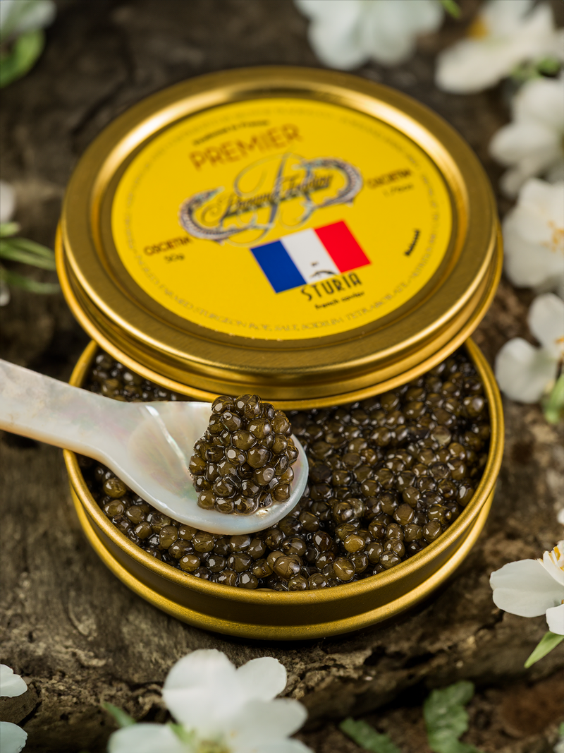 Open gold tin filled with Sturia Premier Osetra caviar and a white mother-of-pearl spoon scooping caviar, placed on rustic wood surrounded by white flowers.