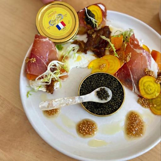 sliced ham and a tin of black caviar on a white plate with sliced radish and mustard seeds.